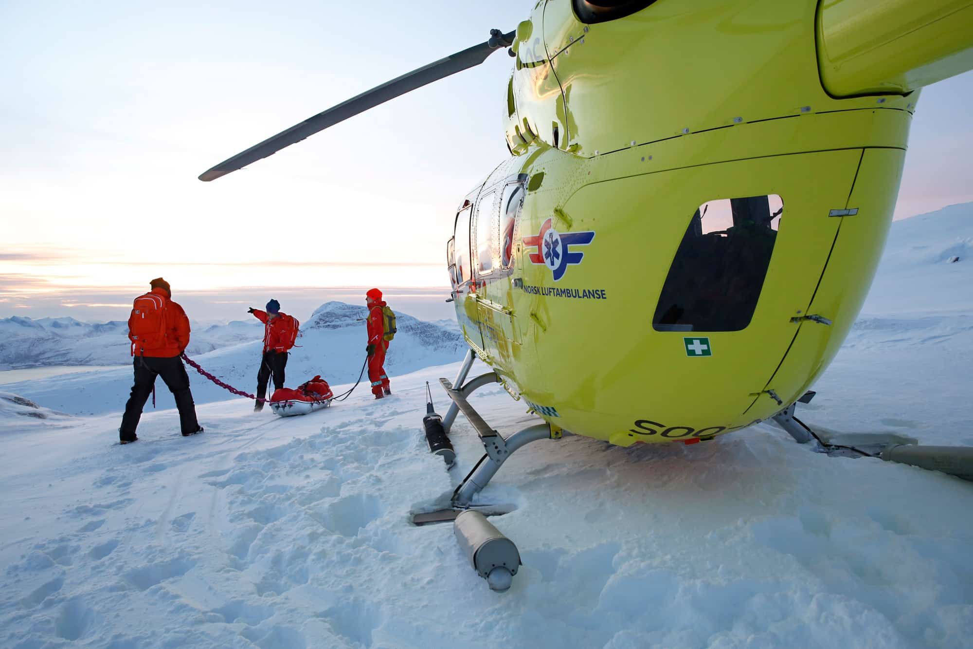 Gult legehelikopter på snødekt fjelltopp. Lege, pilot og redningsmann med utstyr i bakgrunnen.