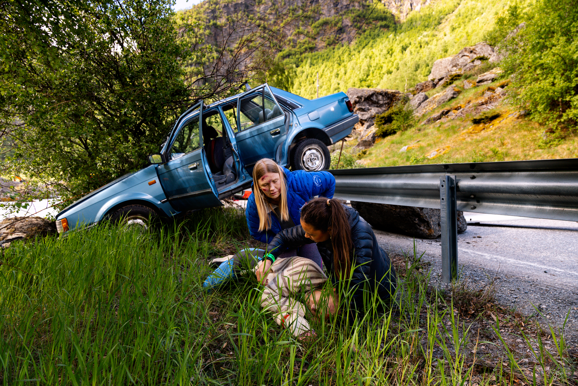 To personer som er lettere skadet sitter bøyd over en tredje hardt skadet person ved veikanten etter en trafikkulykke. Bak dem er en bil som har havnet over autovernet.