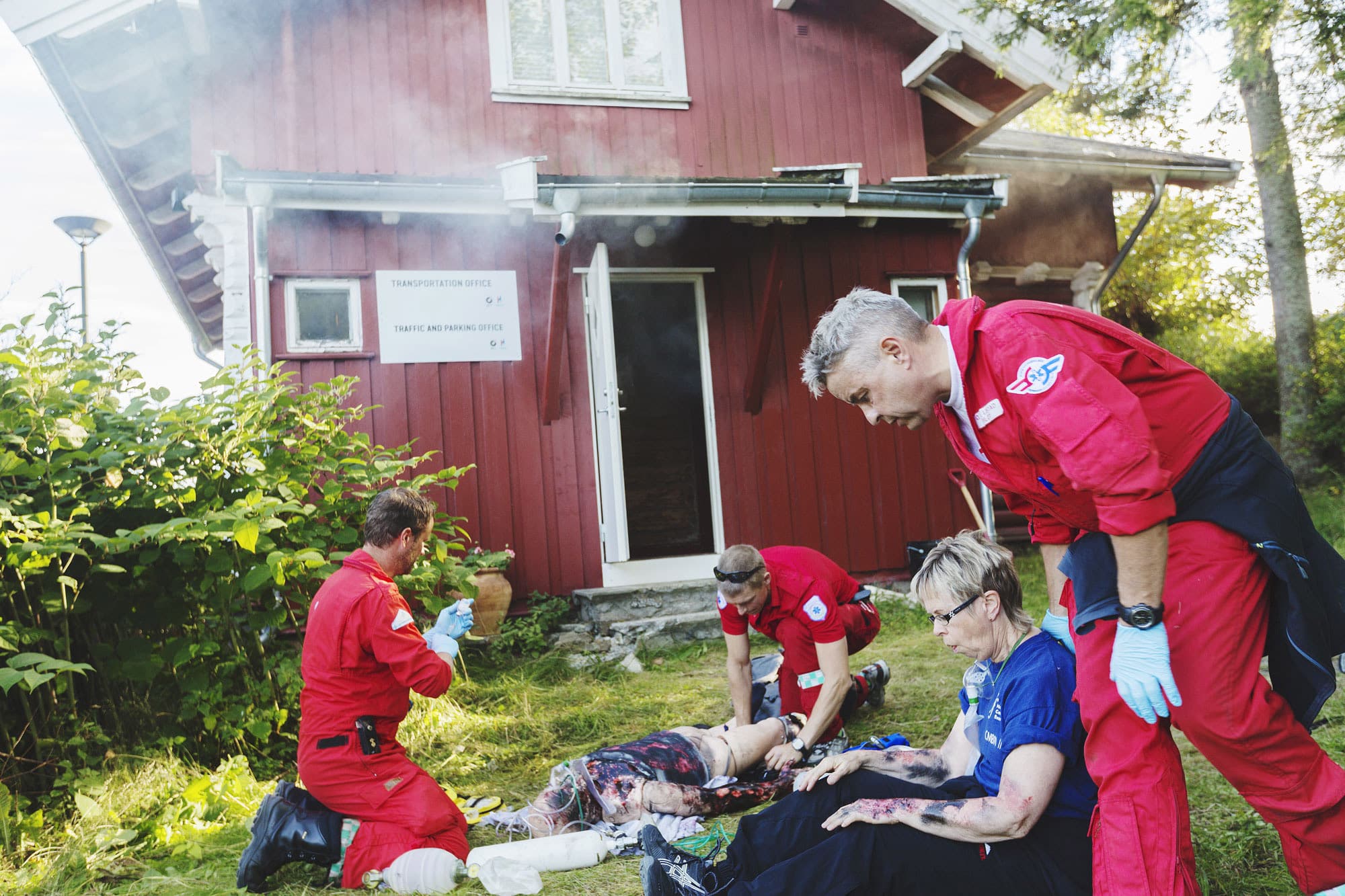 Lege og redningsmann hjelper svært brannskadet pasient under øvelse, mens pilot tar seg av mildere brannskade på en annen. I bakgrunnen et rødt hus det velter røyk ut av.