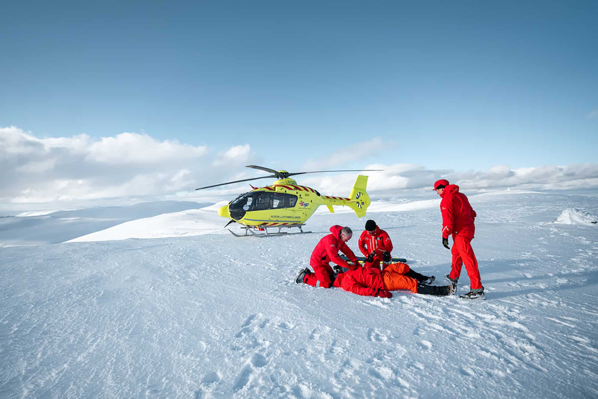 Luftambulansepersonell hjelper pasient på snødekt fjelltopp. Legehelikopter i bakgrunnen.