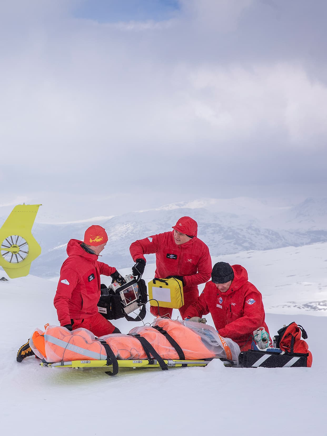 Luftambulansecrew jobber med pasient på båre å snødekt fjelltopp. 