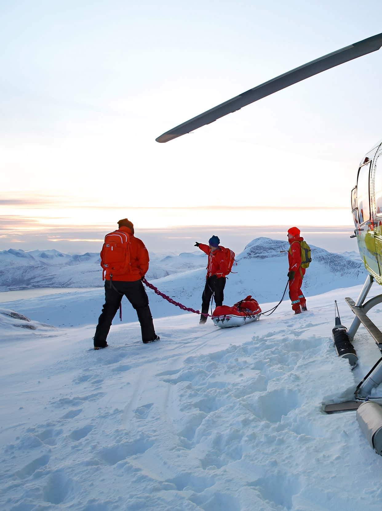 Luftambulansecrew på snødekt fjell foran helikopteret.