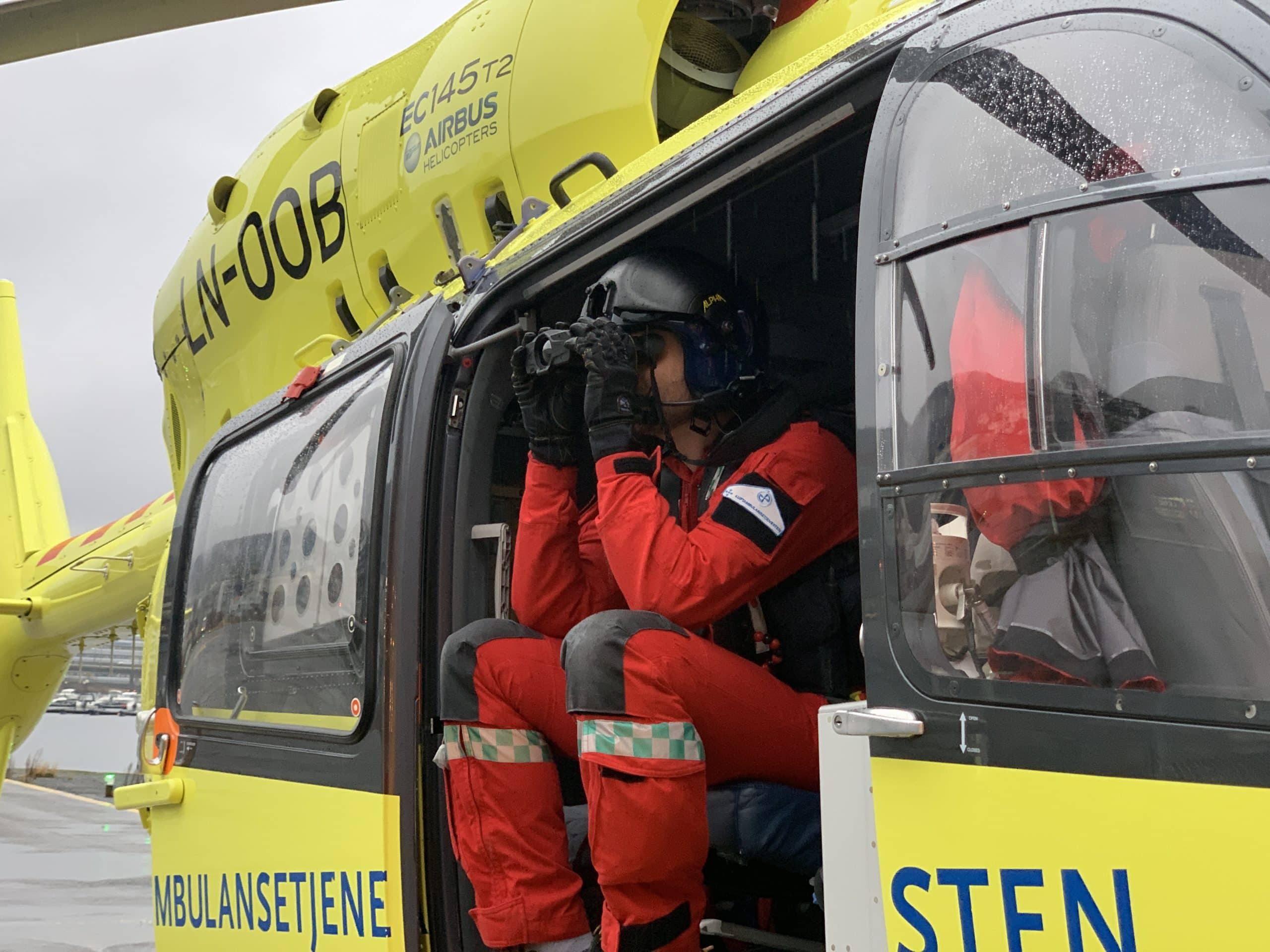 HEMS crew sitting in the door of a helicopter using handheld thermal binoculars. These can be used to detect missing persons who are otherwise unnoticeable in the dark or are difficult to see for other reasons. 