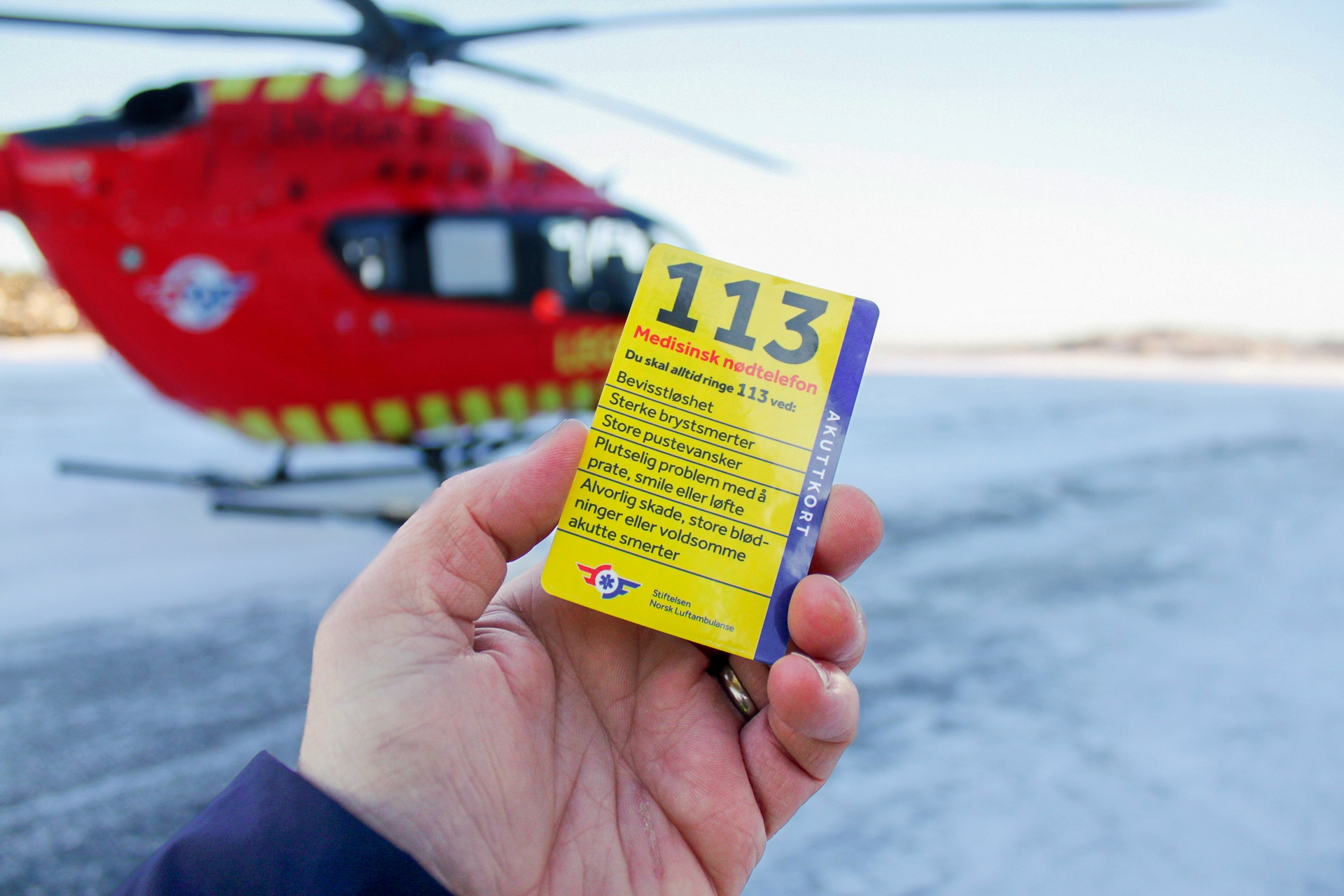 Person holder opp akuttkortet foran et legehelikopter. Akuttkortet forklarer når man bør ringe 113.