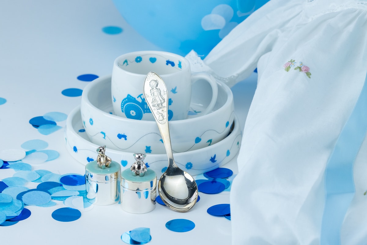 Dåpsgaver til gutt på et bord med blå dekorasjoner, blant annet sølvskje, tallerken og kopp.
