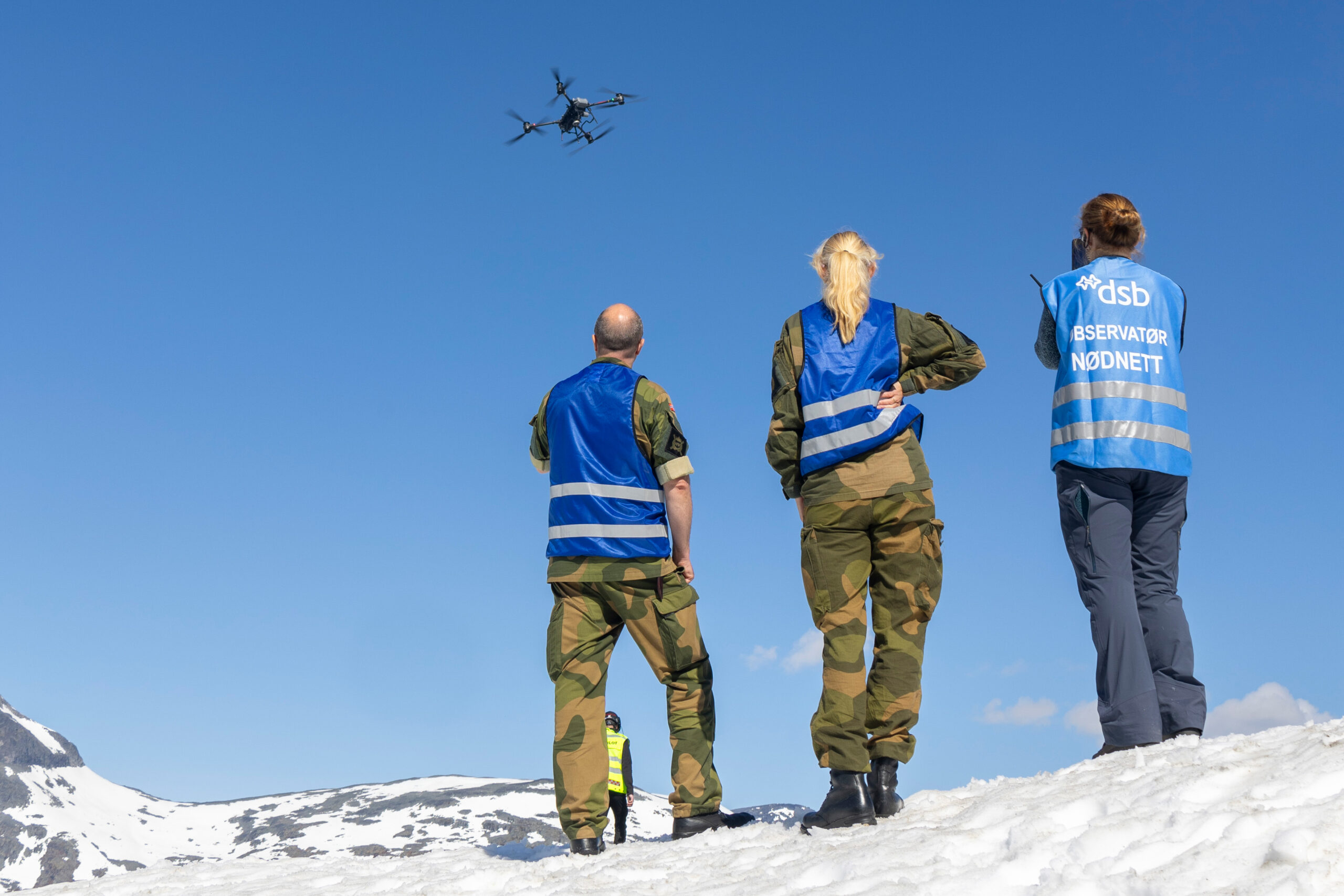 Bilde av tre personer i snølandskap. En drone flyr i lufta.