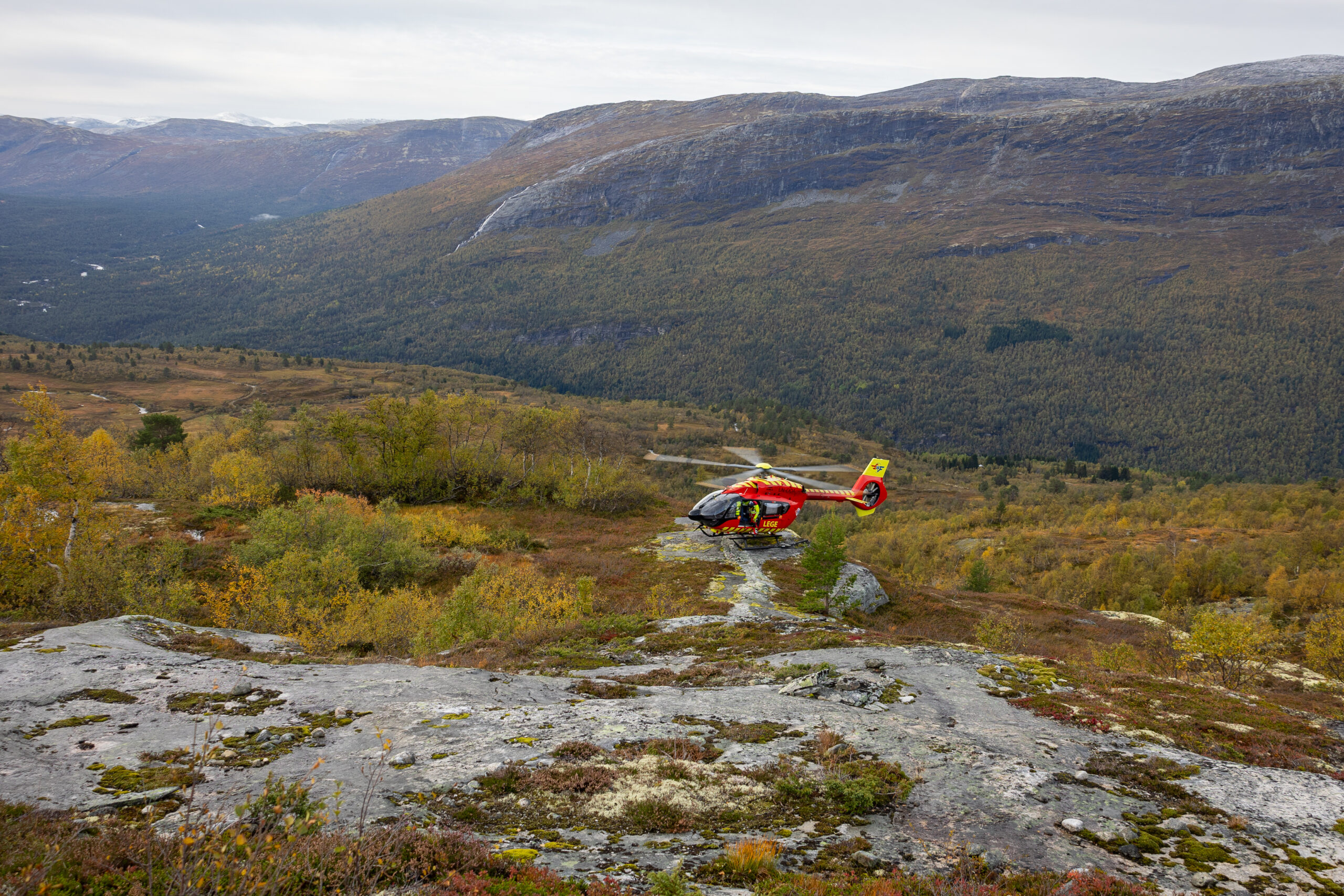 Utviklingshelikopteret flyr over en skog med redningsmannen titter ut av døren