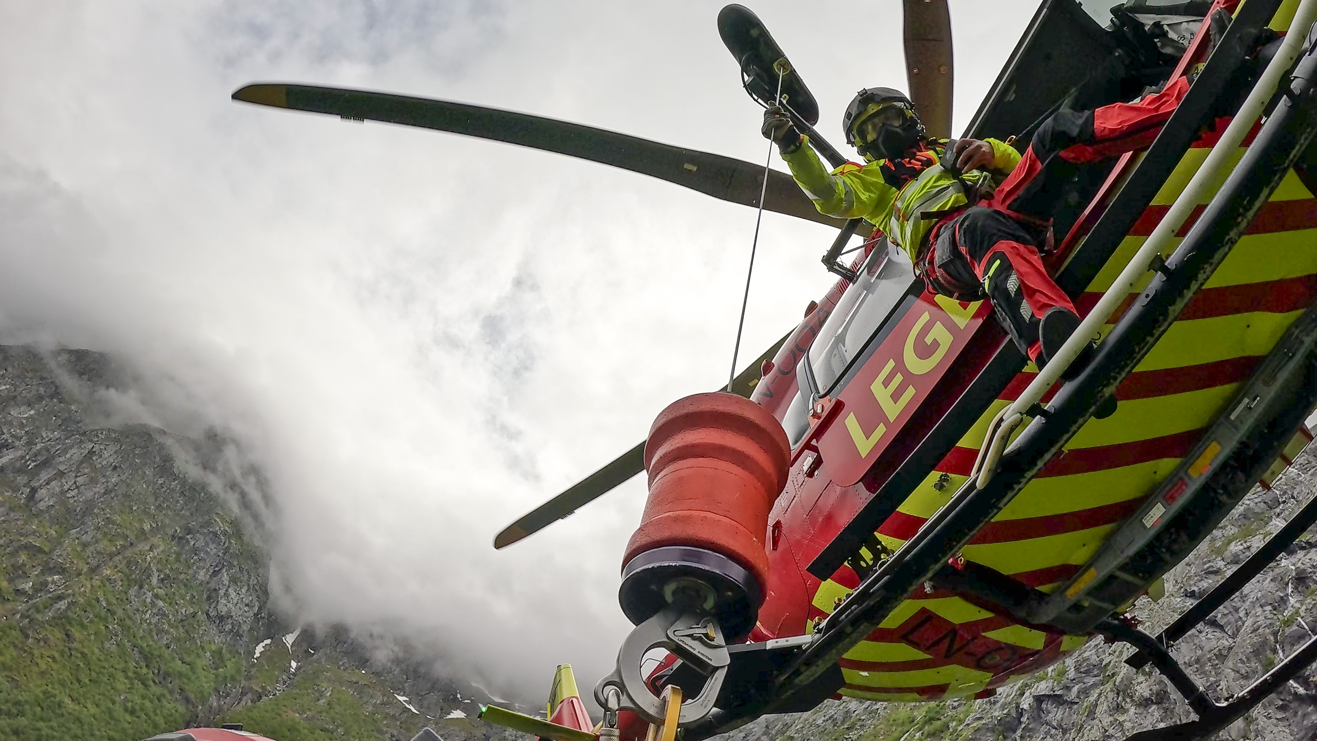Lege står i helikopteret og hjelper redningsmann med å heises ned