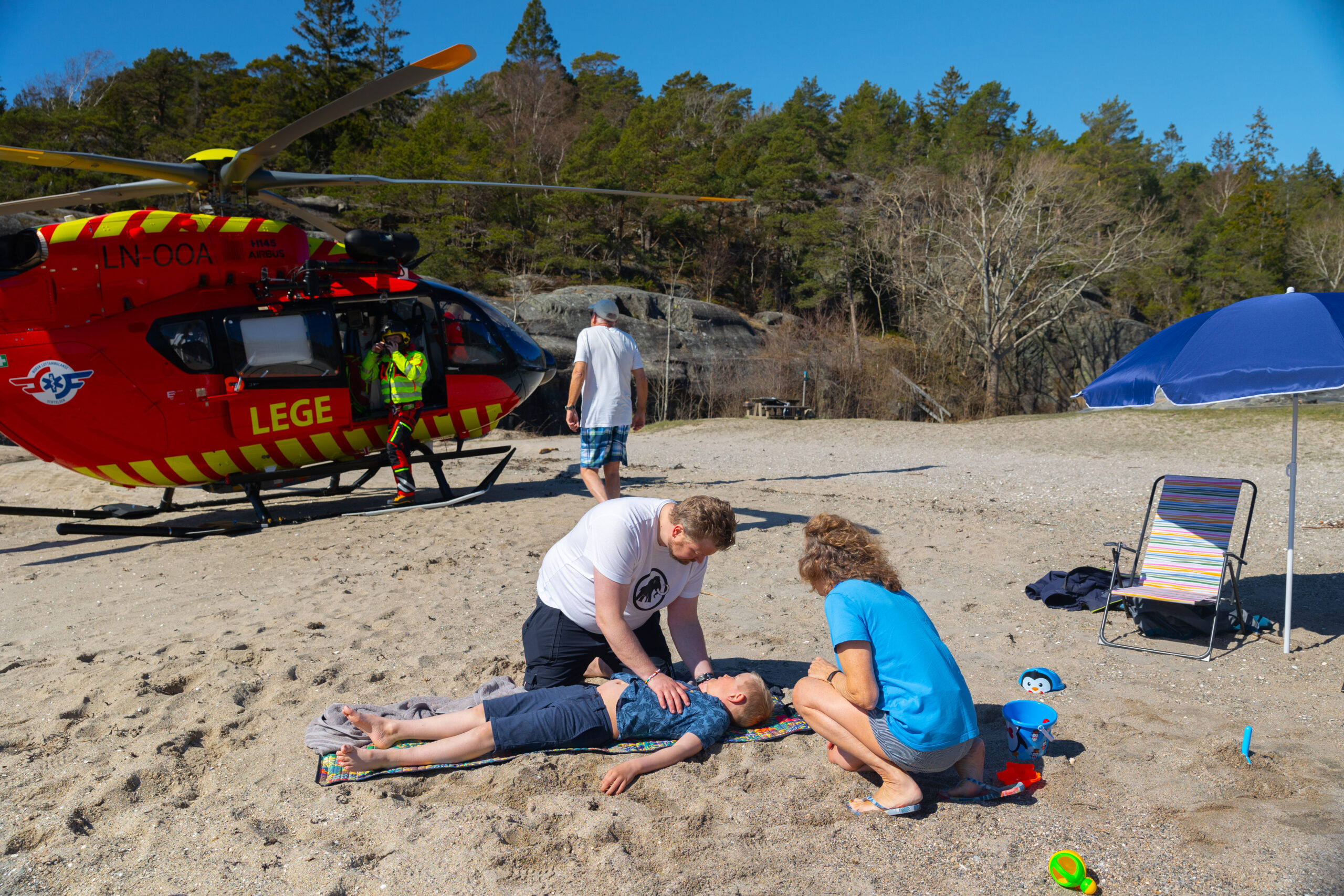 Akutt situasjon med barn, far og besteforeldre på en strand. Legehelikopteret har nettopp landet rett ved. 