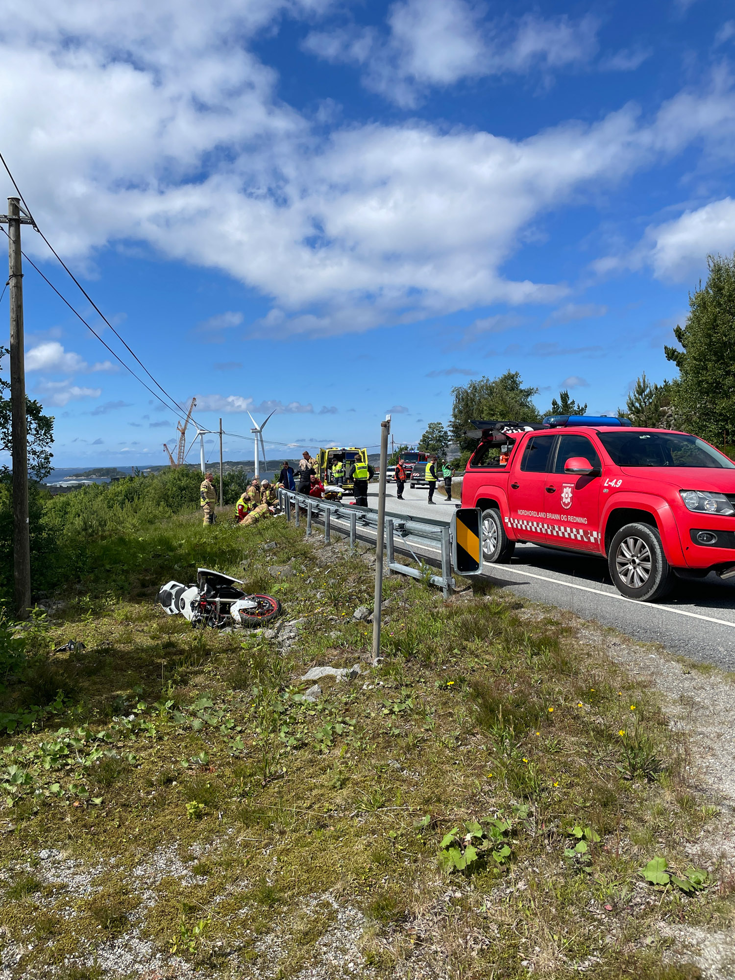 Bilde av skadestedet, der motorsykkelen ligger i grøfta og en utrykningsbil fra brannvesenet står i veikanten.