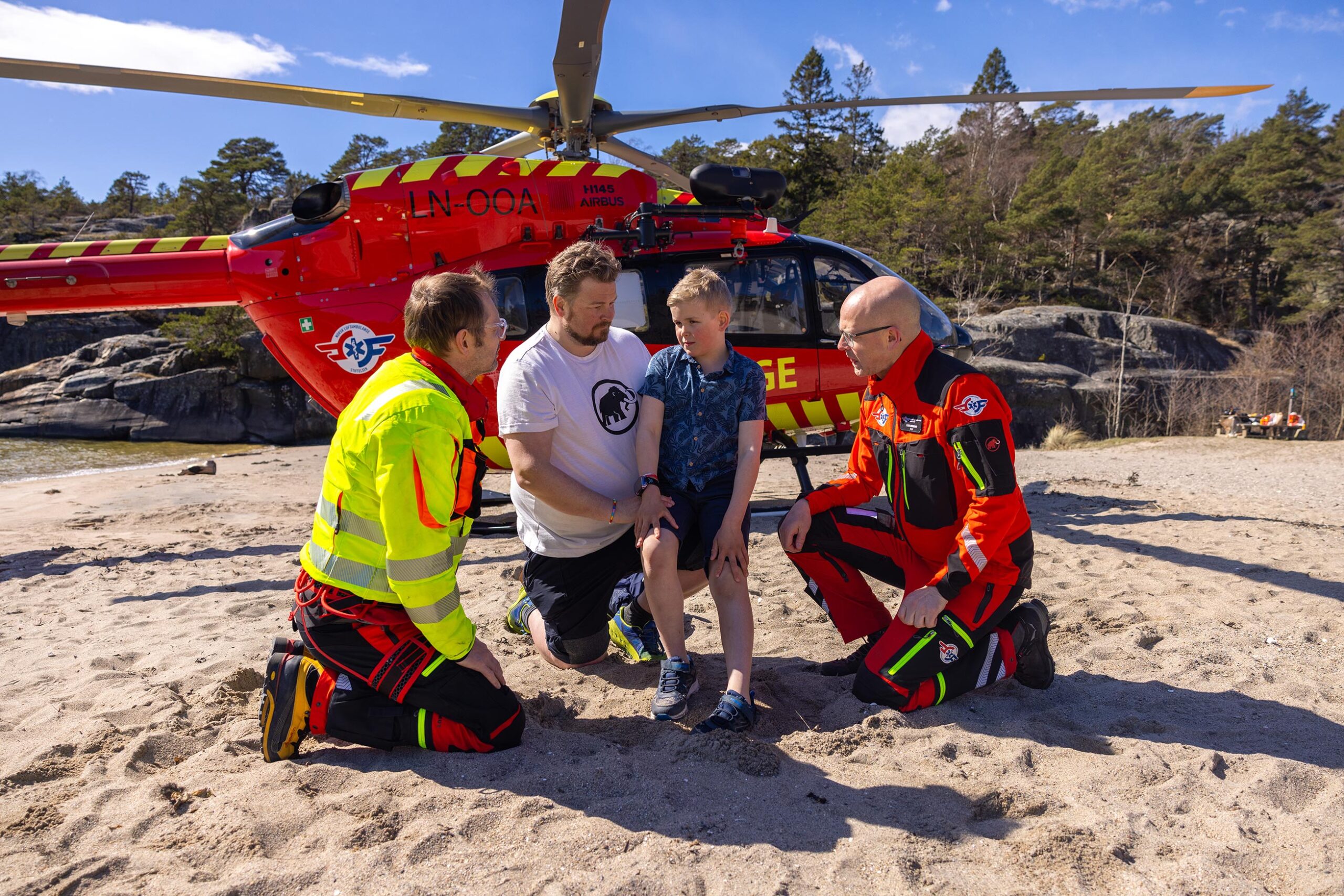 Far og sønn sitter sammen med pilot og luftambulanselege foran helikopteret.